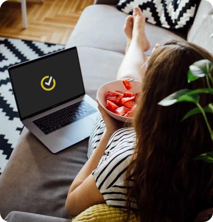 Woman eating watermelon on sofa using laptop launching Norton 360 AntiVirus Plus